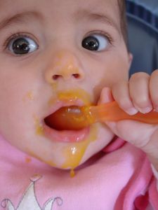 baby holding a spoon and eating strained fruits